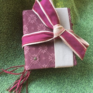 Peut inclure: Un petit livre de couleur bordeaux avec un ruban blanc noué en forme de nœud autour. Le livre a une couverture à motifs et un pompon qui pend en bas.