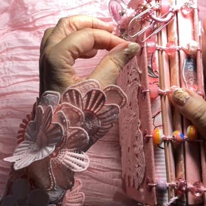 Puede incluir: Un diario rosa con ribete de encaje y cuentas decorativas. El diario está cerrado y tiene una cinta rosa atada alrededor.