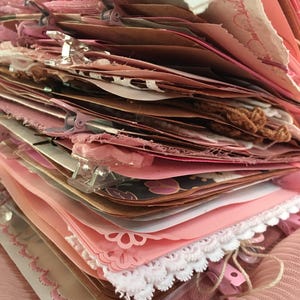 Puede incluir: Una pila de papel rosa y marrón con ribete de encaje blanco y clips de carpeta. El papel está en capas y tiene una estética vintage.