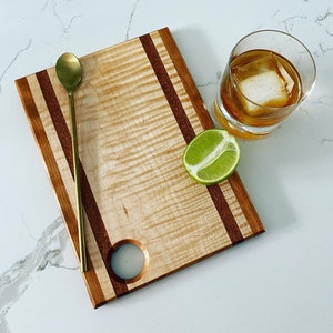 May include: A wooden chopping board with a natural wood finish and two dark brown stripes. The board has a round hole in the centre and a gold spoon on top. A lime wedge and a glass of iced water are on the surface next to the board.