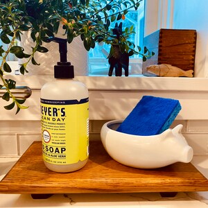 May include: A wooden tray with a white ceramic pig dish holding a blue sponge. A bottle of Meyer's Clean Day dish soap with a yellow label is on the tray. The tray is on a white countertop.