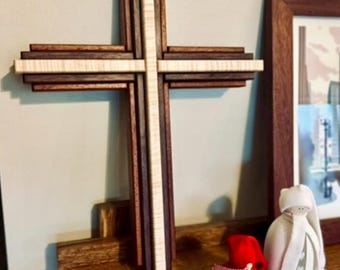 May include: A layered wooden cross with alternating dark and light wood tones. The cross is displayed on a wooden shelf, alongside a red ornament with a white ribbon and a small figurine. The cross is a religious symbol.