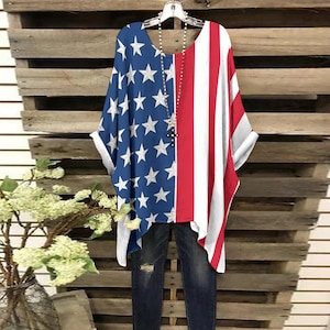May include: A patriotic tunic top featuring a split design: one side with a blue field of white stars, the other with red and white stripes. The top is paired with dark blue jeans. A necklace with a cross pendant hangs in the center.