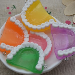 May include: Five colourful plastic teeth models with white enamel and silver rings. The teeth are arranged in a circle on a white plate. The colours are orange, yellow, green, pink, and purple.