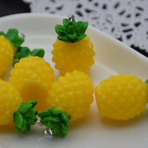Puede incluir: Seis abalorios de piña amarillos con tapas verdes y lazos plateados. Los abalorios de piña están en un plato blanco.