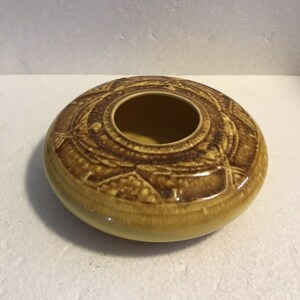 May include: A yellow ceramic ashtray with a brown geometric design. The ashtray has a round shape and a center hole.