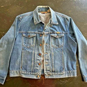 May include: A medium-wash blue denim jacket featuring a classic collar, long sleeves with buttoned cuffs, and a front button closure. The jacket includes two chest pockets with buttoned flaps and two vertical side pockets, finished with copper-toned buttons.