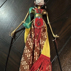 May include: An Indonesian Wayang Golek puppet. The puppet has a painted face, gold-coloured arms, and is dressed in a red, brown, and yellow patterned costume with red fringe. The puppet is held by black rods.