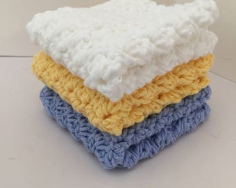May include: Three crocheted dishcloths stacked on top of each other. The top dishcloth is white, the middle dishcloth is yellow, and the bottom dishcloth is blue.