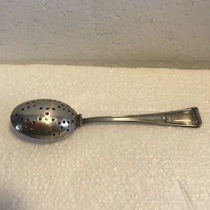 May include: A silver tea infuser with a perforated oval bowl and a long handle. The handle has a decorative rectangular end. The infuser is designed for steeping loose leaf tea. The tea infuser is a kitchen utensil.