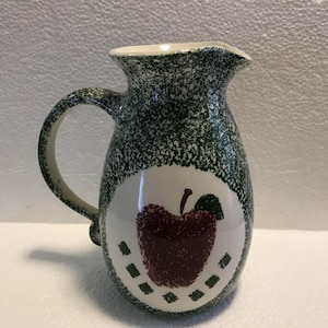 May include: A ceramic pitcher featuring a dark red apple illustration on a white circular background, surrounded by a green speckled sponge-painted pattern. The pitcher has a curved handle and a pouring spout, with small green squares below the apple.