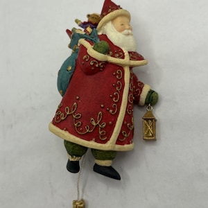 May include: A Santa Claus figurine wearing a red coat with gold scrollwork and cream trim, green trousers, and black boots. Santa carries a blue sack filled with toys on the back and holds a small gold lantern. A gold gift charm hangs from a string.