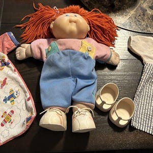 May include: A vintage Cabbage Patch Kids doll with red yarn hair, wearing blue overalls and white shoes. Included are a pink and white airplane-themed sleep sack and a striped overall outfit with a white shirt.