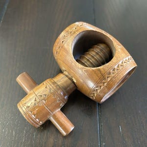 Puede incluir: Un cascanueces de madera con mecanismo de tornillo. El cascanueces es de madera marrón claro y presenta patrones geométricos tallados alrededor de la abertura y en el mango. El mango tiene forma de cruz.