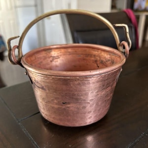 May include: A small, hammered copper bucket with a brass handle. The bucket has a rounded shape and a slightly textured surface. The handle is arched and attached to the bucket with small, curved brackets. The copper has a warm, reddish-brown hue.
