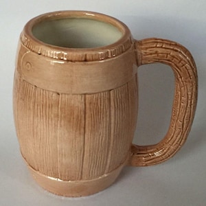 May include: A ceramic mug with a barrel design. The mug is light brown with vertical wood grain texture and two horizontal bands. The handle is shaped like a tree branch. The interior is a light cream color.