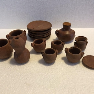 May include: A miniature terracotta kitchen set featuring several small brown clay pots with handles, a stack of round flat plates, and a small jug. The collection consists of various unglazed earthenware vessels and dishes arranged together.