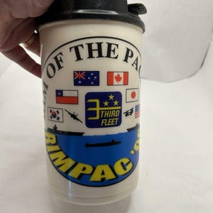 May include: Cream-colored plastic travel mug with a black lid. The mug features the text "RIMPAC" in yellow and blue, along with flags and the words "OF THE PACIFIC". The mug has a black flip-top lid.