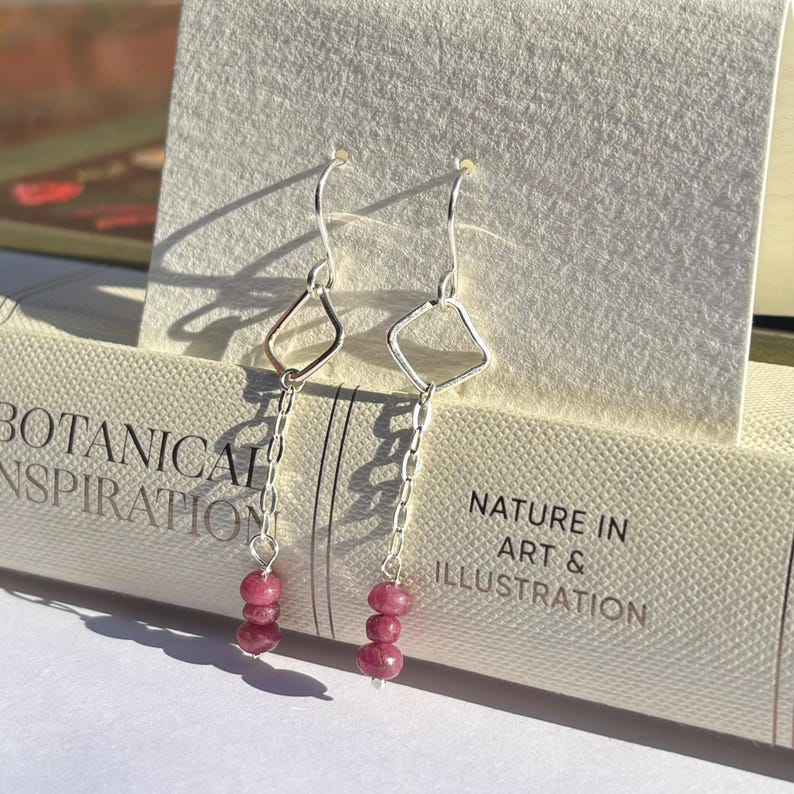 May include: A pair of silver dangle earrings with a geometric diamond shape and a chain of three pink beads. The earrings are displayed on a book with the text "BOTANICAL INSPIRATION" and "NATURE IN ART & ILLUSTRATION".