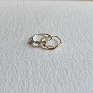 May include: Two thin metal rings with an irregular, wavy, and textured surface. One ring is silver-toned and the other is gold-toned. The rings are linked together, showcasing a delicate, organic, and uneven band design with small raised details.