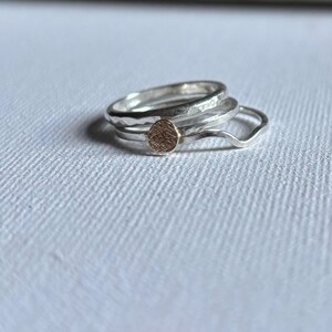 May include: A set of three stacked rings consisting of two silver bands with a hammered texture and one thin, wavy silver band. The centre ring features a small, circular, textured gold-toned accent piece attached to the front of the band.