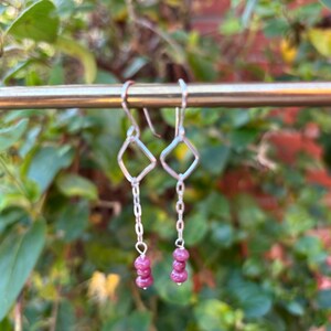 May include: Silver dangle earrings with a geometric diamond shape and a chain that leads to three small, round, pink gemstones. The earrings are hanging in front of a blurred green and red background.