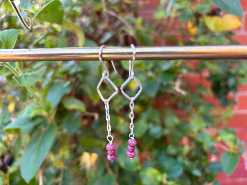 May include: Silver dangle earrings with a geometric diamond shape and a chain that leads to three small, round, pink gemstones. The earrings are hanging in front of a blurred green and red background.