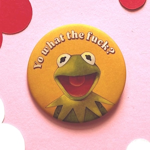 Könnte beinhalten: Ein gelber Knopf mit einem Cartoon-Bild von Kermit dem Frosch und dem Text "Yo what the fuck?". Der Knopf ist vor einem rosa Hintergrund mit roten und weißen Papierformen platziert.