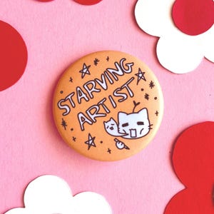 Könnte beinhalten: Orangener Knopf mit dem Text "STARVING ARTIST" und einer Cartoon-Illustration von zwei Katzen. Der Knopf hat einen weißen Umriss und schwarzen Text.