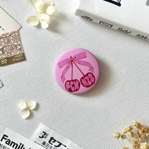 Könnte beinhalten: Ein runder, rosafarbener Ansteckbutton mit einer grafischen Illustration von zwei Kirschen, die mit einer rosa Schleife zusammengebunden sind. Der Text FCK NZS ist in einer fetten, dunkelrosa Schrift auf den Kirschen mittig abgebildet.