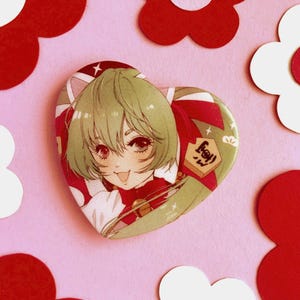 Könnte beinhalten: Eine herzförmige Anstecknadel mit einer Comicfigur mit grünen Haaren, die ein rot-weißes Outfit trägt. Die Figur lächelt und hat eine rote Schleife auf dem Kopf. Die Anstecknadel hat eine glitzernde Oberfläche und den Text "Happy!" in Japanisch.