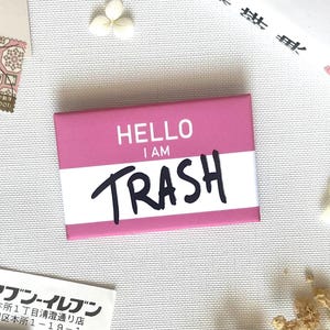 Könnte beinhalten: Ein rechteckiger, rosa-weißer Namensschild-Pin mit dem Text HELLO I AM in weißen Großbuchstaben auf rosa Hintergrund und dem Wort TRASH in großer, fetter, schwarzer Handschrift auf dem weißen Mittelstreifen.