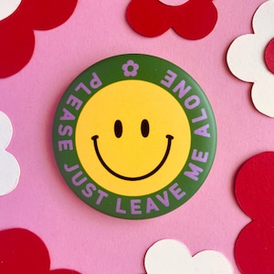 Könnte beinhalten: Ein grüner Knopf mit einem gelben Smiley und dem Text "Please Just Leave Me Alone" in lila Schrift.