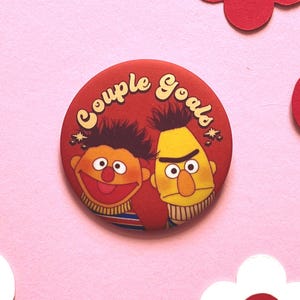 Puede incluir: Un botón redondo rojo con la inscripción "Couple Goals" en amarillo estilizado. El botón presenta ilustraciones de dibujos animados de dos personajes, uno naranja y otro amarillo, con rostros expresivos. Elementos decorativos rodean el botón.