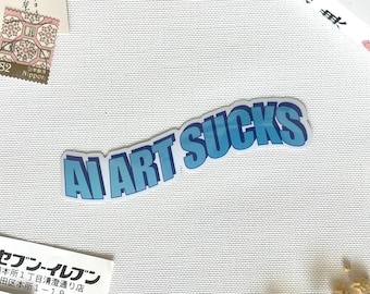 May include: A die-cut sticker featuring the text AI ART SUCKS in bold, light blue capital letters with a dark blue drop shadow. The sticker has a white border and is positioned against a textured white background with other paper ephemera visible.