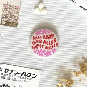 Op de afbeelding: Een ronde button met een lichtroze achtergrond en rode tekst die luidt Ich kann das alles nicht meer, omringd door kleine roze bloemillustraties. Het ontwerp is gecentreerd op een getextureerd wit oppervlak met diverse papieren items.
