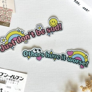 Könnte beinhalten: Zwei gestanzte Aufkleber mit glitzerndem Text und bunten Grafiken. Der obere Aufkleber zeigt den rosa Glitzertext Just don't be sad! mit einem gelben Smiley und Regenbogen. Der untere Aufkleber hat den blauen Glitzertext Others have it worse! mit Regenbogen und Herzen.
