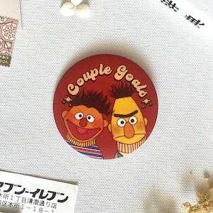 Op de afbeelding: Een ronde sticker met een rode achtergrond met de personages Bert en Ernie uit Sesamstraat. De tekst "Couple Goals" is in een gebogen, crèmekleurig lettertype boven de personages gedrukt, die gestreepte shirts dragen.