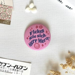 Könnte beinhalten: Ein runder, rosafarbener Ansteckbutton mit dunkelblauem Text, der Ficken Sie sich, Herr Merz! lautet. Das Design enthält kleine rote Blumen- und Sternsymbole sowie ein einfaches rotes Smiley-Gesicht, das unten auf dem Button zentriert ist.