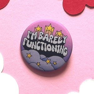 Könnte beinhalten: Ein runder Button mit einem rosa-lila Farbverlauf. Der Button zeigt den Text "I'M BARELY FUNCTIONING" in Weiß und Schwarz, mit gelben Sternen und Wolken. Ein lustiges und skurriles Accessoire.