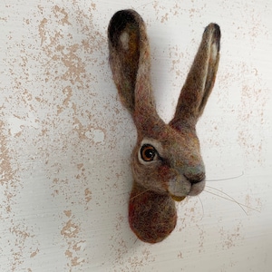 May include: A brown and gray felt sculpture of a rabbit's head with long ears. The rabbit has a brown nose and a single brown eye.