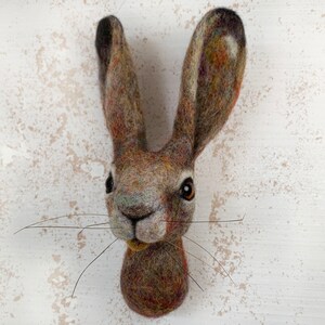 Puede incluir: Una escultura de fieltro marr&oacute;n y gris de la cabeza de una liebre con largos bigotes. La liebre tiene ojos marrones y una nariz blanca.