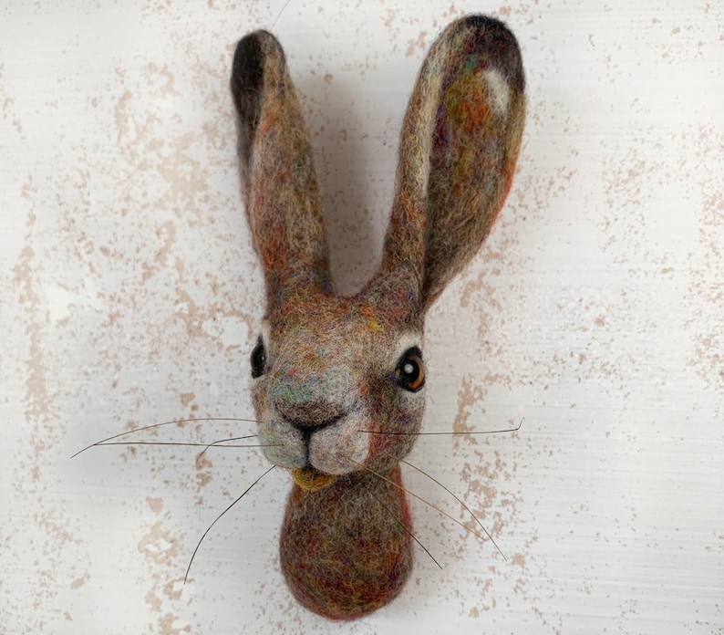 Puede incluir: Una escultura de fieltro marr&oacute;n y gris de la cabeza de una liebre con largos bigotes. La liebre tiene ojos marrones y una nariz blanca.