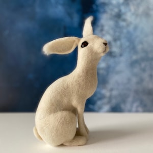 May include: A white felt rabbit figurine sitting on a white surface. The rabbit has a long, fluffy tail and is looking to the right.