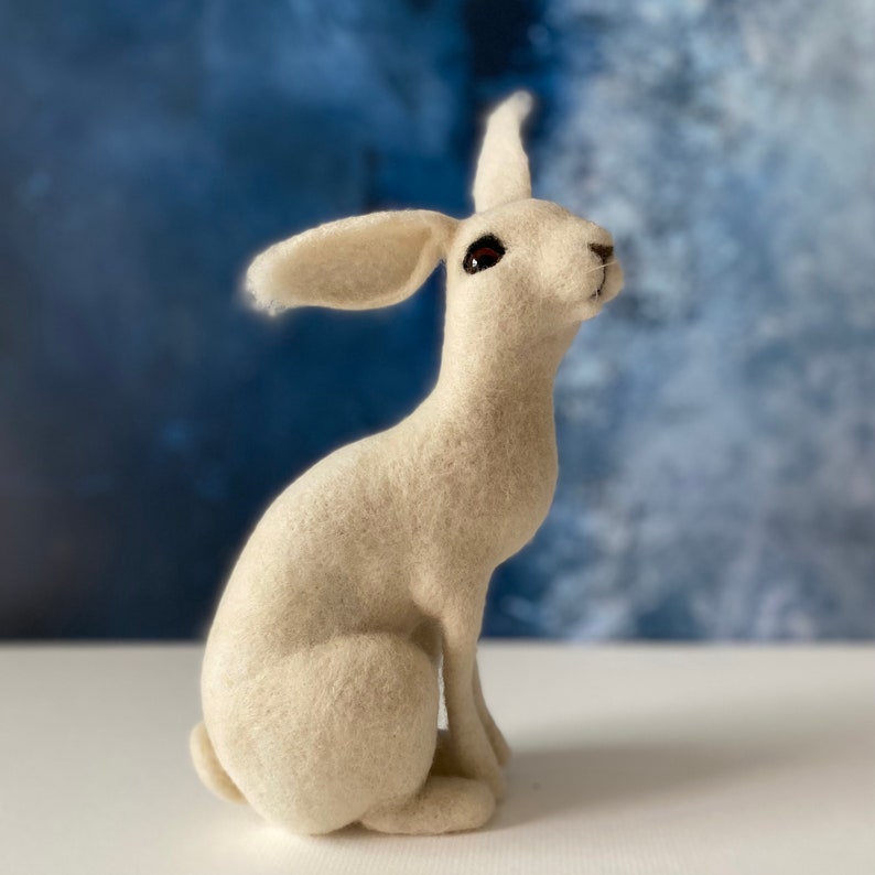 May include: A white felt rabbit figurine sitting on a white surface. The rabbit has a long, fluffy tail and is looking to the right.