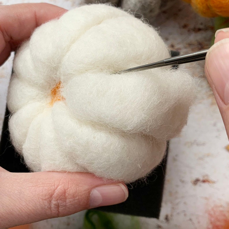 May include: A white felted pumpkin with a small orange center is being felted with a needle tool.