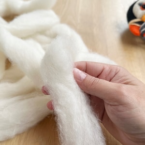 May include: A close-up of a hand holding a strand of white wool roving. The wool is soft and fluffy, and it is perfect for felting or spinning.
