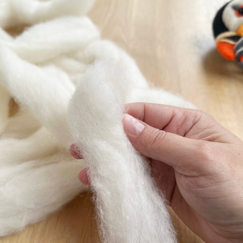 May include: A close-up of a hand holding a strand of white wool roving. The wool is soft and fluffy, and it is perfect for felting or spinning.