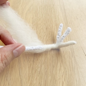 May include: A white fluffy yarn is wrapped around a white pipe cleaner with two prongs at the end. The pipe cleaner is bent to form a shape resembling a bird's head.