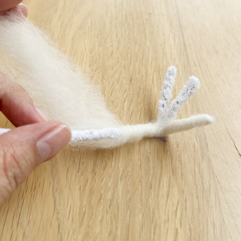 May include: A white fluffy yarn is wrapped around a white pipe cleaner with two prongs at the end. The pipe cleaner is bent to form a shape resembling a bird's head.